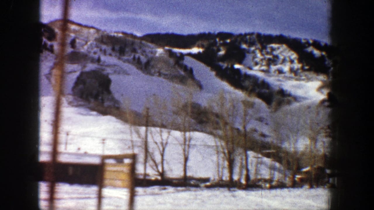 Snow-covered mountain slope with trees, utility poles, and small structures in the foreground.