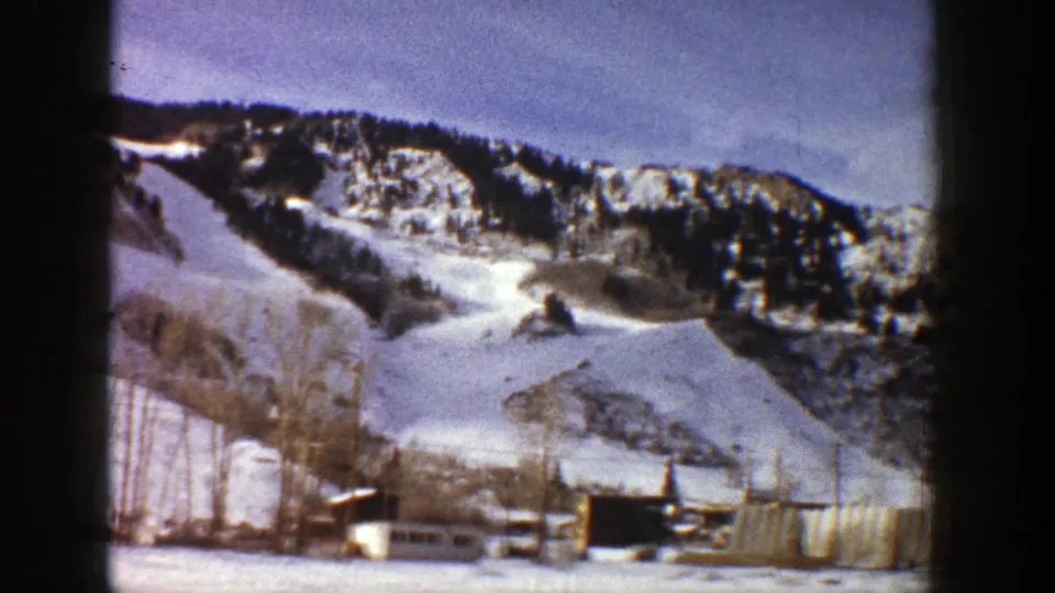 1961 Aspen 8mm frame showing a snow-covered hillside, dark evergreen ridge, bare trees, and structures along the valley edge.