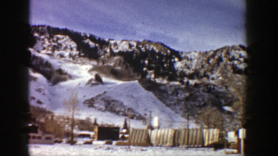 Aspen, Colorado winter frame with snow-covered mountain terrain, bare trees, utility poles, and low foreground structures on 8mm film.