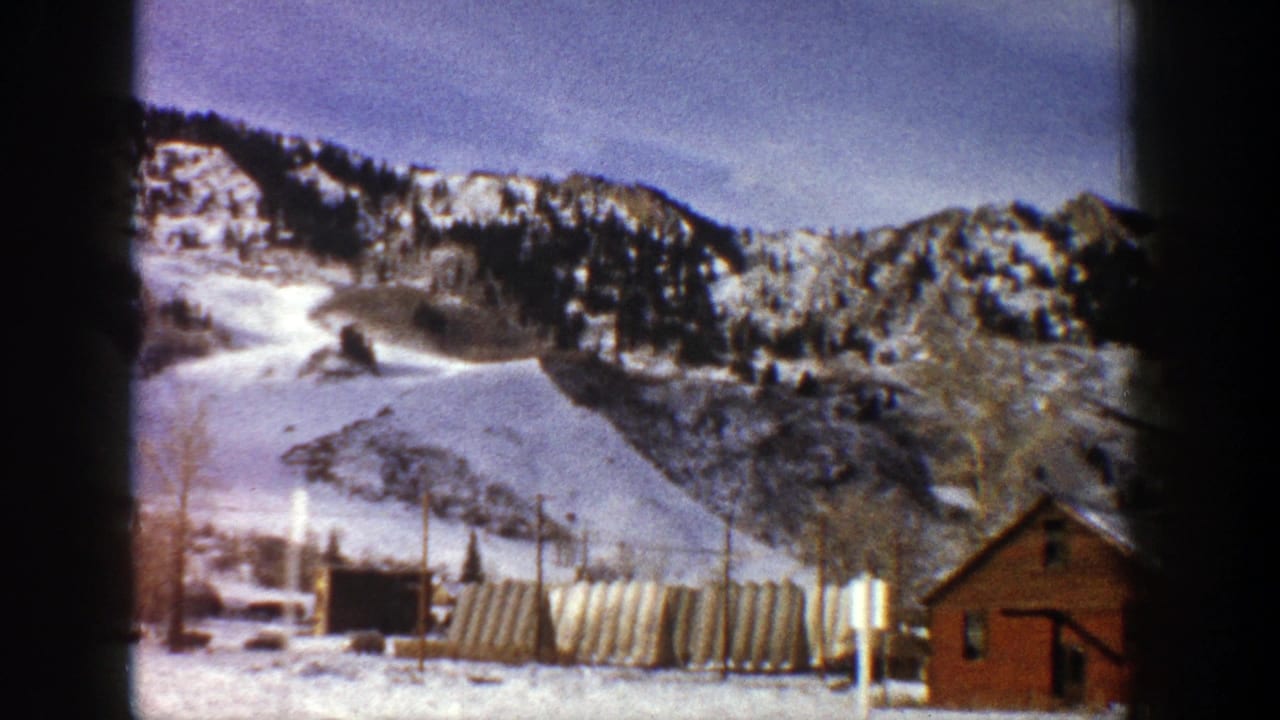 1961 Aspen, Colorado home-movie frame showing snow-covered mountains, resort buildings, fencing, and a red cabin.