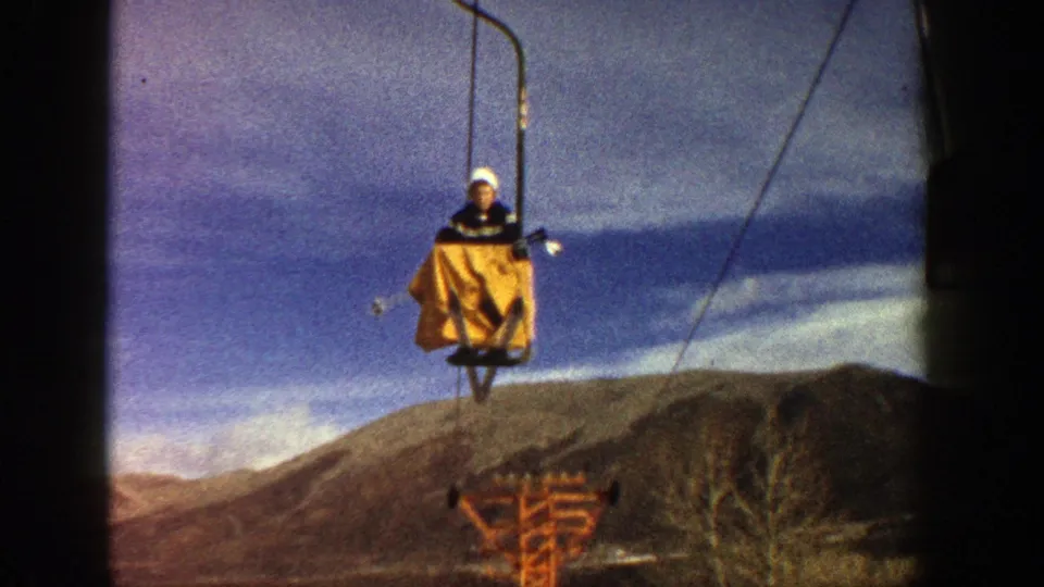 A skier in a yellow jacket and white hat rides a chairlift against blue sky and Colorado mountain terrain.