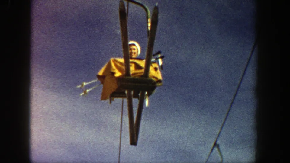 Low-angle 8mm frame of the chairlift rider in yellow clothing, with skis and lift hardware silhouetted against blue sky.