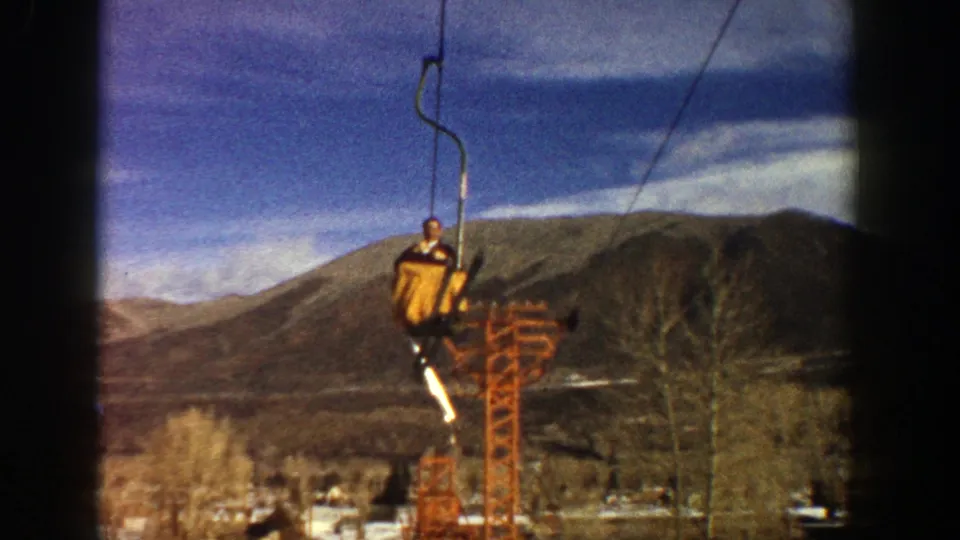 1961 Aspen chairlift scene with a skier in yellow passing a tower above bare trees and mountain slopes.