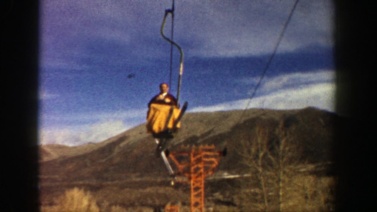 In Aspen, Colorado, 1961, a man in yellow ski pants rides an open chairlift against distant mountains and blue sky.