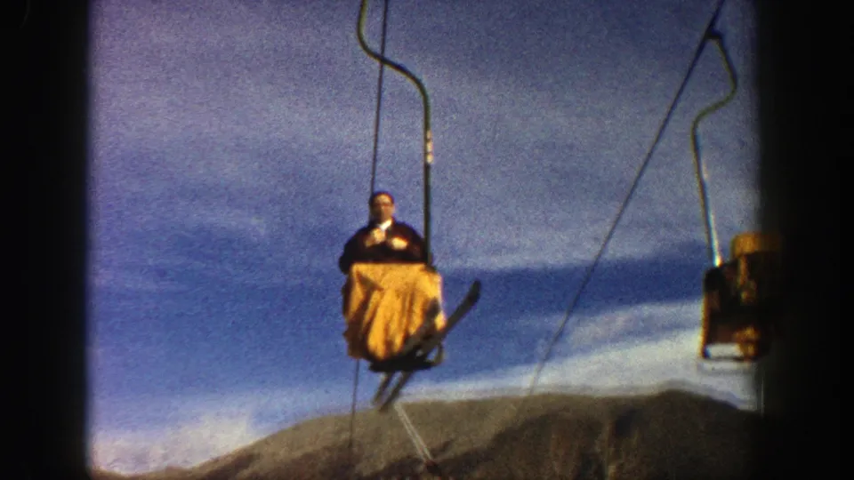 A 1961 Aspen chairlift rider moves above the lift area, with a red support structure, mountains, and blue sky visible.