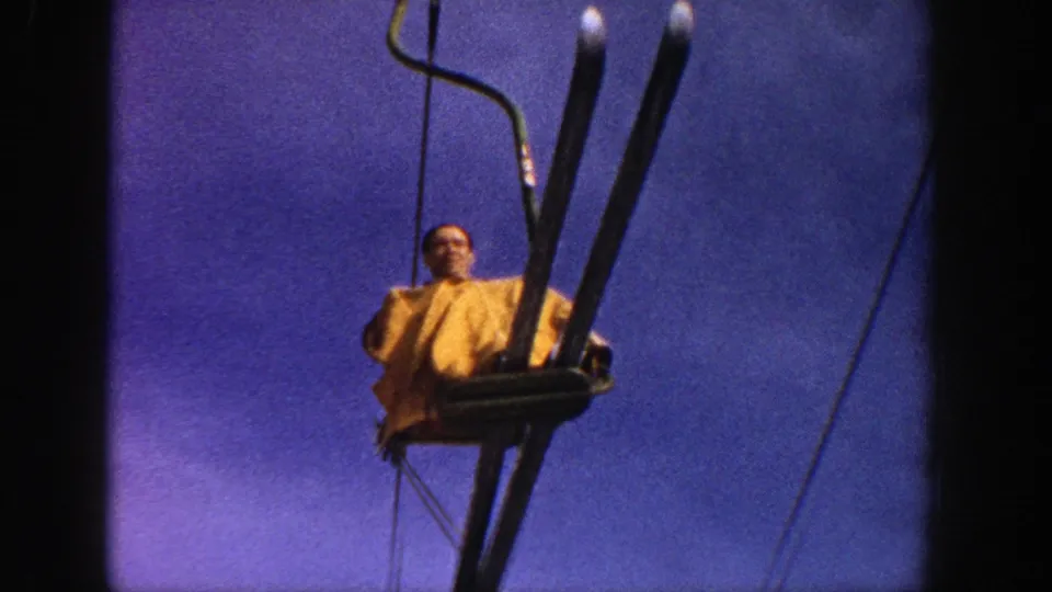 In the Aspen ski-resort footage, the rider holds upright skis while seated on the open chairlift against a blue sky.