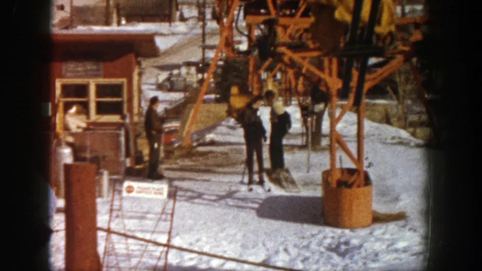 The Aspen lift base shows snow, a red building, chairlift machinery, and several people standing near the loading area in 1961.
