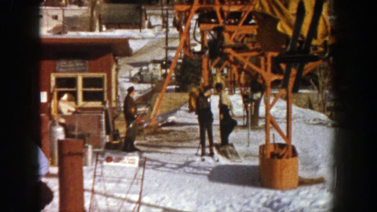 1961 Aspen ski resort scene with skiers and staff near a wooden ticket booth, orange cable car structure, and sunlit snow.