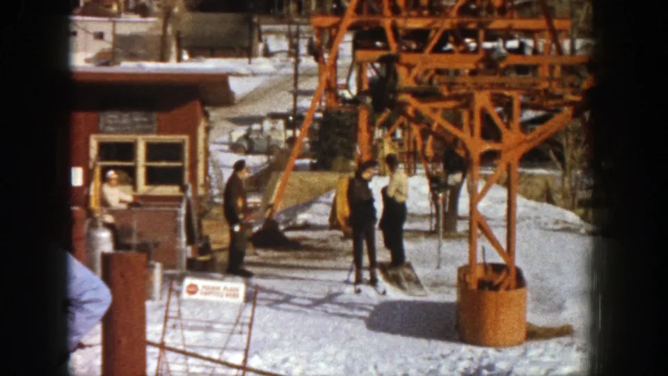 At the Aspen cable car station, staff and skiers stand near the lift structure while a person sits inside the booth.
