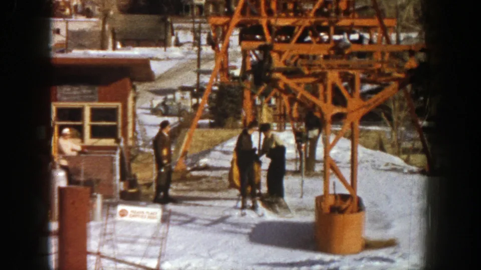 Skiers and resort staff stand in conversation at the 1961 Aspen station, framed by orange lift supports and snow.