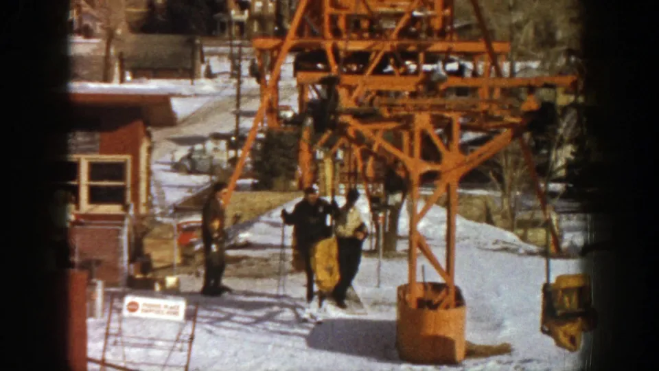At the Aspen ski station, two people in winter clothing walk across the snow as another stands beside the booth and lift structure.