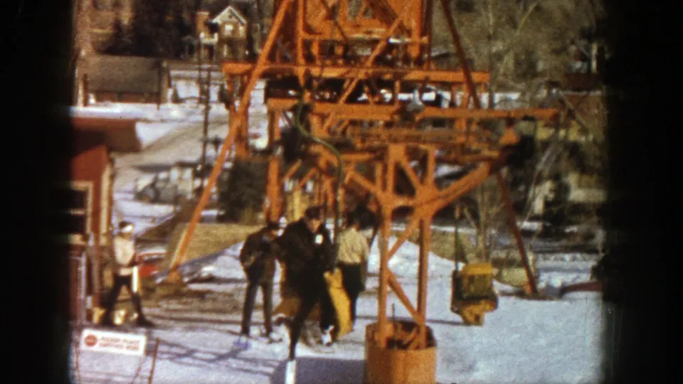 A small group of skiers moves through the snowy lift area beneath an orange chairlift tower in 1961 Aspen.
