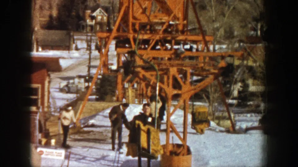 At an Aspen ski lift in 1961, a skier stands beside a yellow chair with the orange tower and snow behind.