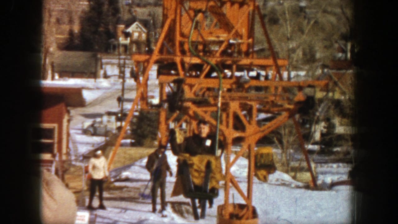 1961 Aspen home-movie frame showing a rider in a brown coat on an orange ski lift with skiers waiting on snow below.