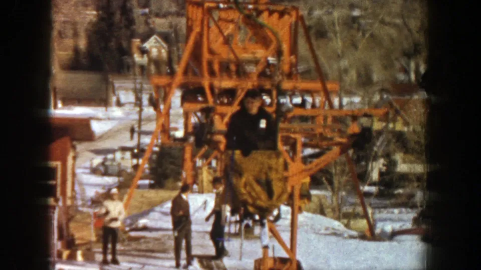 The Aspen ski lift approaches the camera, with a rider in a brown coat seated above a snowy base area.
