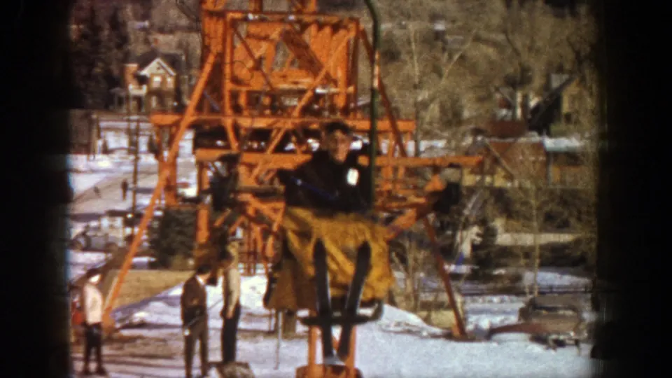 A 1961 Aspen ski-area view centers the lift rider against an orange tower and snowy ground with people nearby.