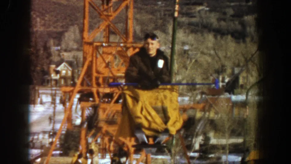 Close 1961 Aspen frame of a male lift rider in dark and brown winter clothing, with the orange ski-lift tower behind him.
