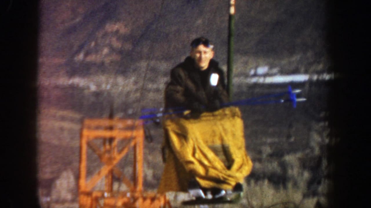 Man seated on a yellow-covered ski-lift chair near an orange lift structure in snow.