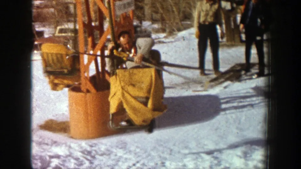 A man rides a yellow-covered chair near an orange ski-lift tower in the 1961 Aspen home-movie footage.