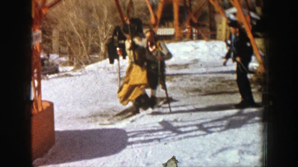 In the Aspen 1961 footage, the man appears on skis near the orange lift structure while other people stand on the snowy slope.