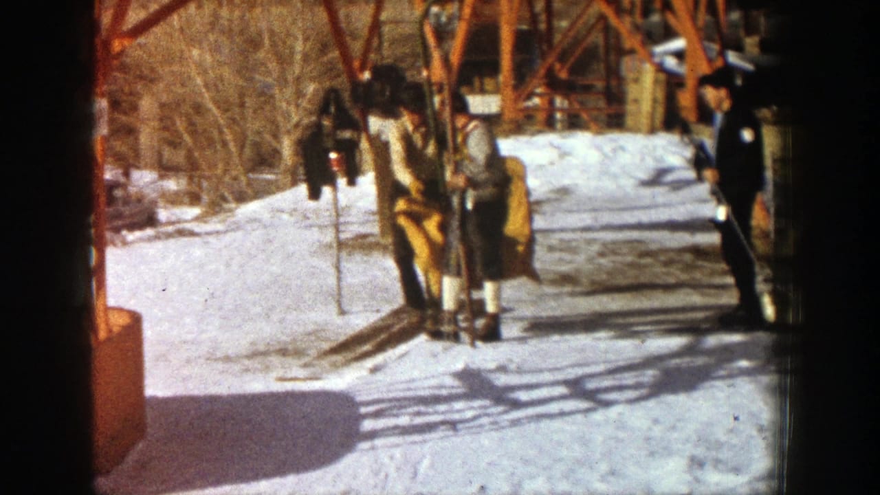 Two men on skis stand near a chairlift while another man waits beside the lift structure.