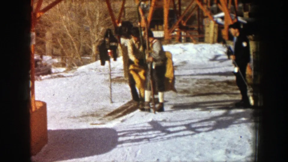 In Aspen, Colorado in 1961, two adult skiers stand on snow near a chairlift as a third man watches beside the metal support area.