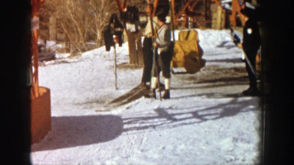 Two adult skiers in winter clothing stand at the Aspen chairlift loading area with skis and poles visible in the snow.