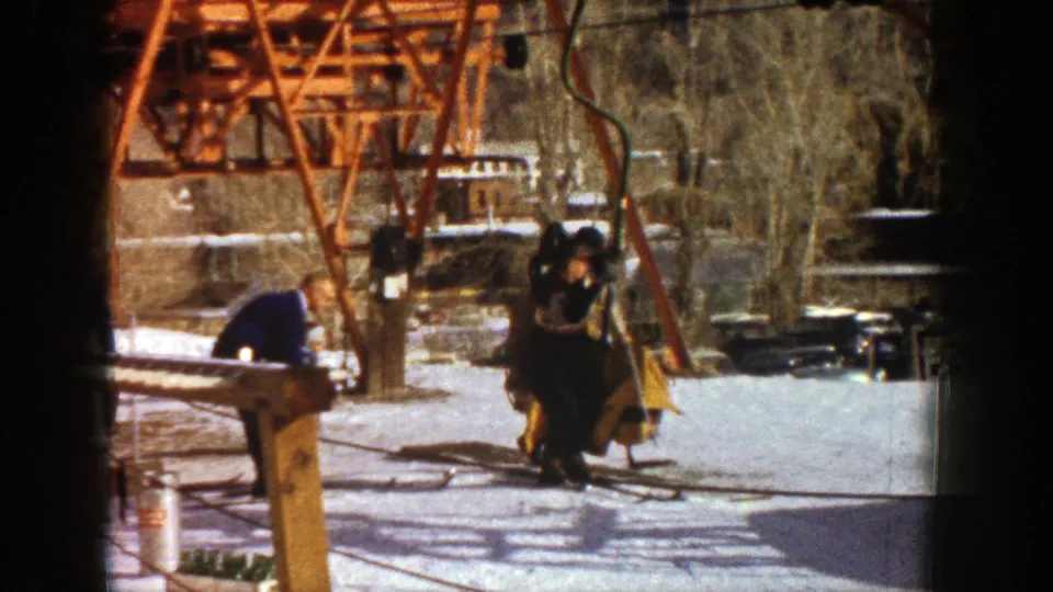 At the 1961 Aspen ski lift, one man bends forward while another skier is low beside the chair, suggesting assistance during boarding.