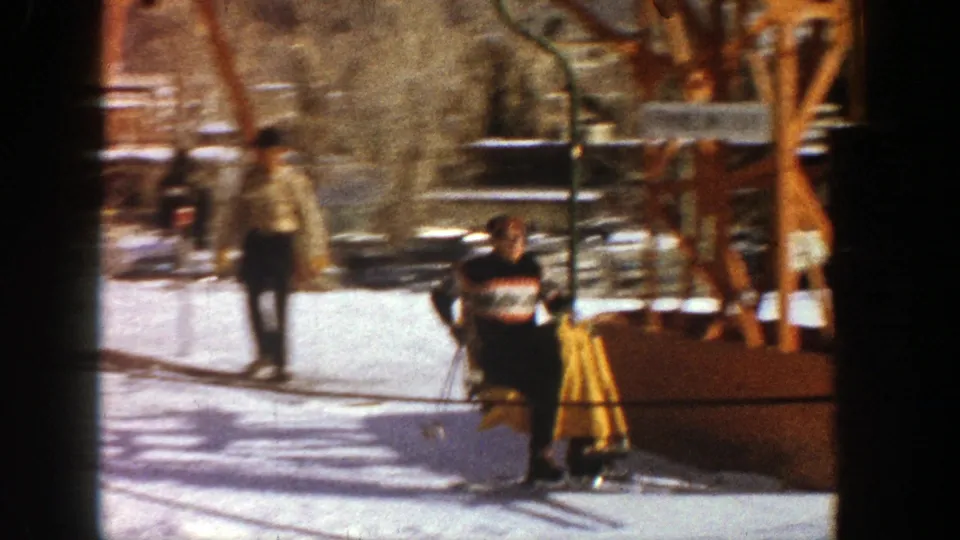 A skier in dark winter clothing is seen on or beside the chairlift at Aspen, with another man crossing the snowy resort area behind him.