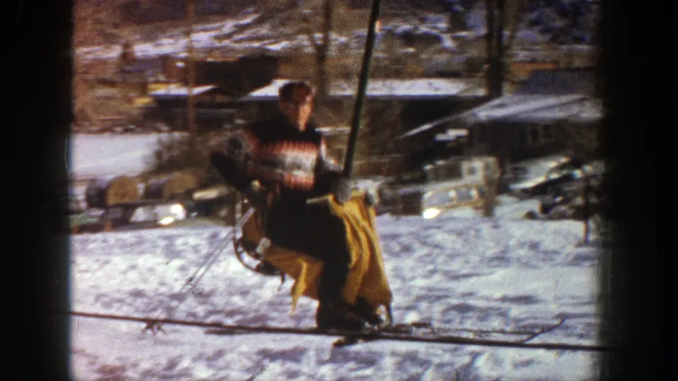 A man in a patterned ski sweater rides a chairlift at Aspen in 1961, passing snowy ground, parked cars, and resort buildings.