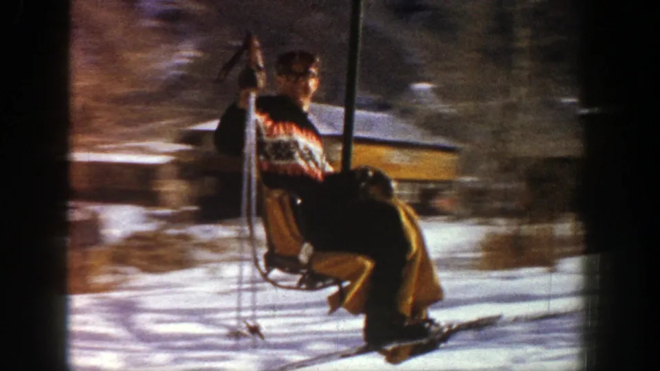 Side view of a man on an Aspen chairlift in 1961, wearing ski clothing and holding poles as the snowy background blurs.