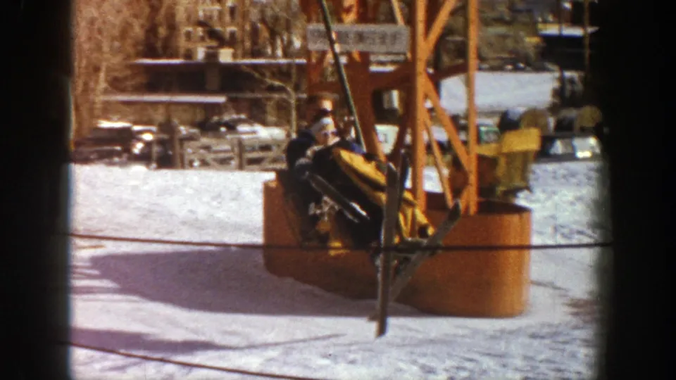The skier passes close to a wooden lift structure at the Aspen resort, with snow, cables, and parked cars visible behind him.