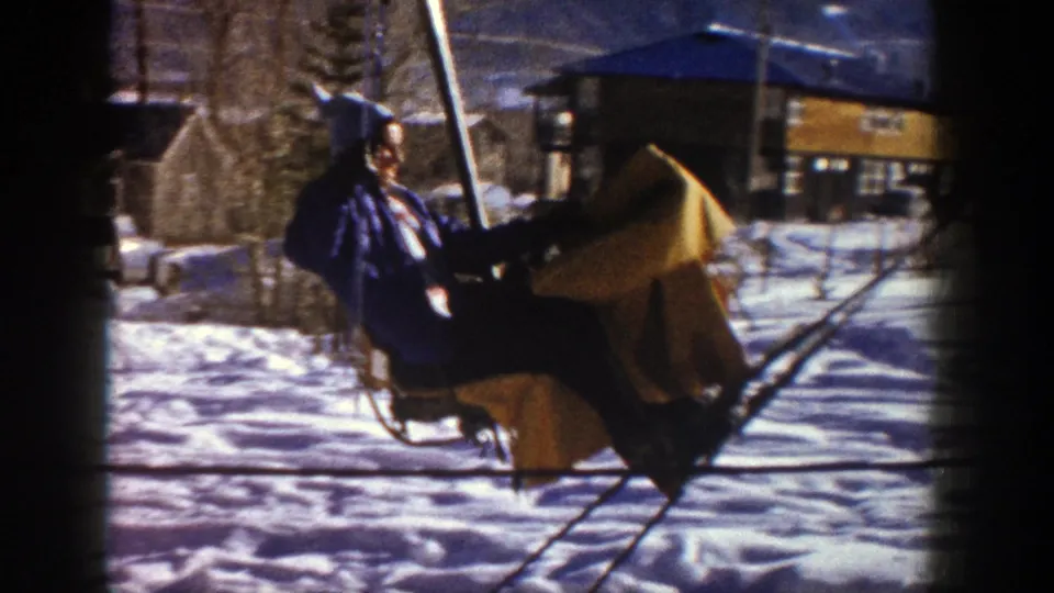 Young woman in blue winter clothing rides a ski lift at Aspen, Colorado, with snow and lodge buildings behind her.