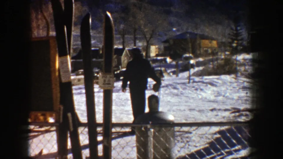 Aspen ski-area frame with a skier moving across snow near upright skis, fencing, trees, and resort buildings.