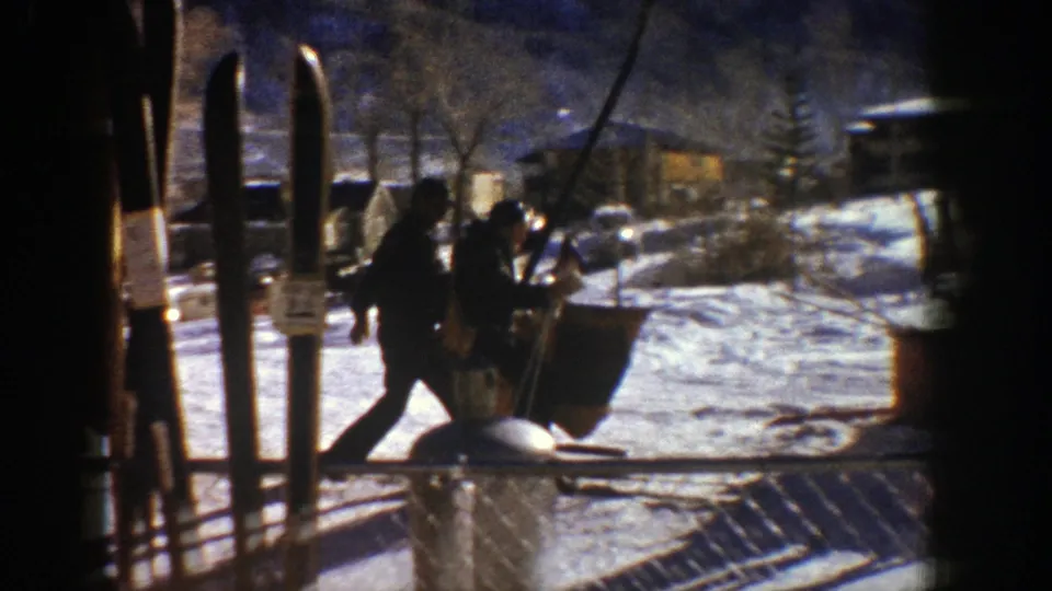 People pass through the Aspen ski base area near upright skis, snow paths, trees, and a lift structure.