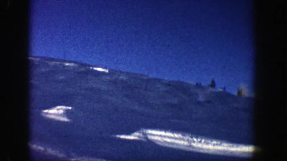 From a chairlift or elevated viewpoint in Aspen, the camera looks across a blue-toned snowy mountain slope with small trees and bright patches of reflected light.