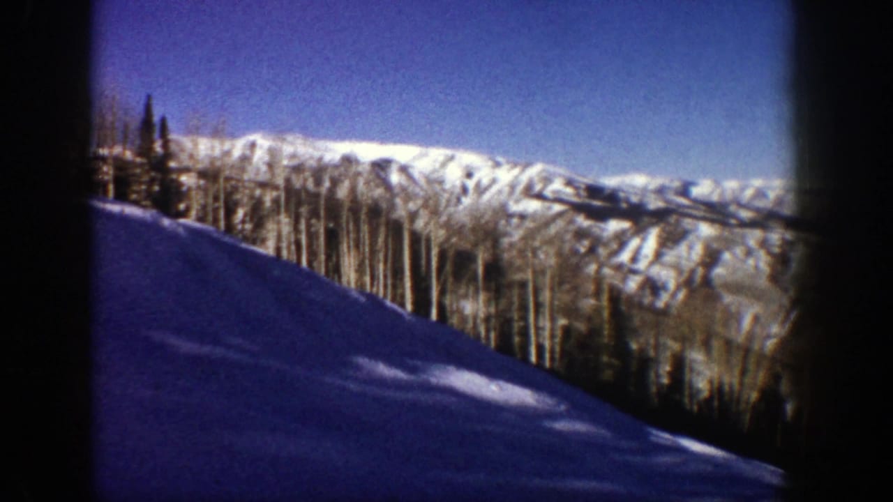 Snowy downhill slope with forest and distant mountain ridges under a blue sky.