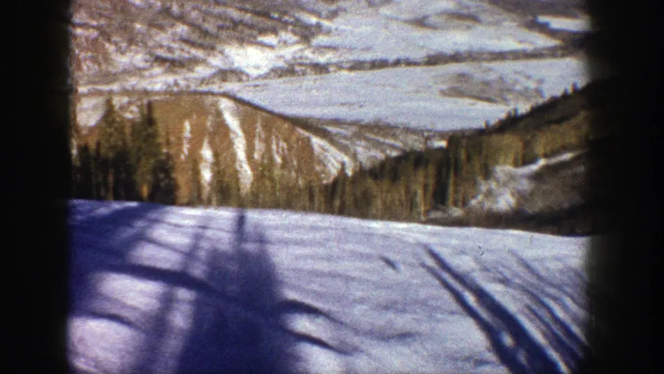1961 Aspen home-movie frame showing a snow-covered slope, tree line, and distant mountain ridges through dark foreground framing.