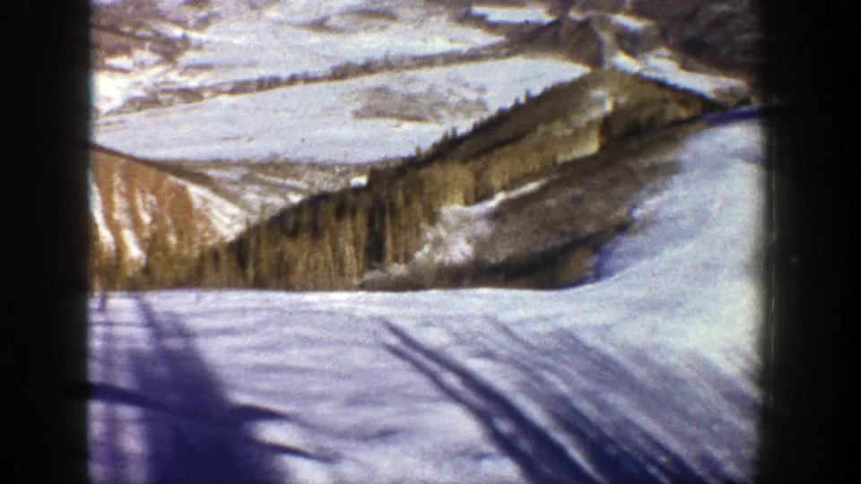 1961 Aspen frame with snow tracks crossing a bright slope, evergreen forest, and brown-white mountain ridges beyond.