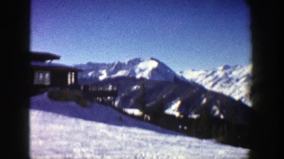 1961 Aspen frame revealing a dark lodge-like mountain building set against snow-covered peaks and a clear blue sky.