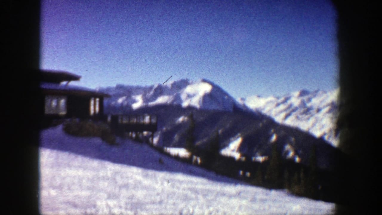 1961 Aspen 8mm frame showing snow-covered mountains, blue sky, and a slope-side structure at the left edge.
