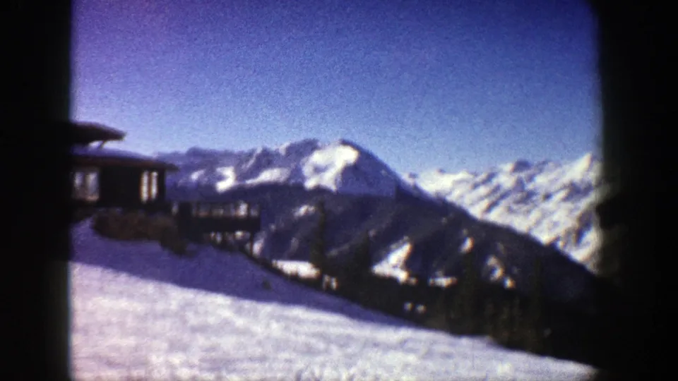 1961 Aspen home-movie frame with a wooden structure at left and snowy Rocky Mountain ridges beyond.