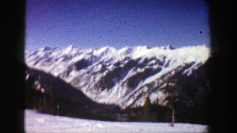 Likely Aspen-area 1961 8mm view of snow-bright mountain peaks, blue sky, dark lower terrain, and visible film gate edges.