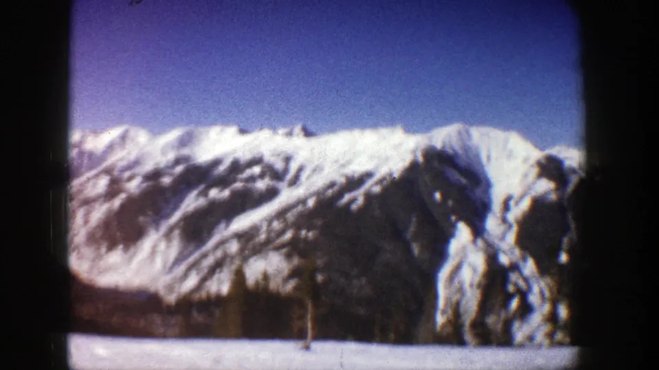 Likely Aspen-area 1961 8mm frame showing a snow-covered ridgeline, steep mountain face, dark trees, and blue sky.