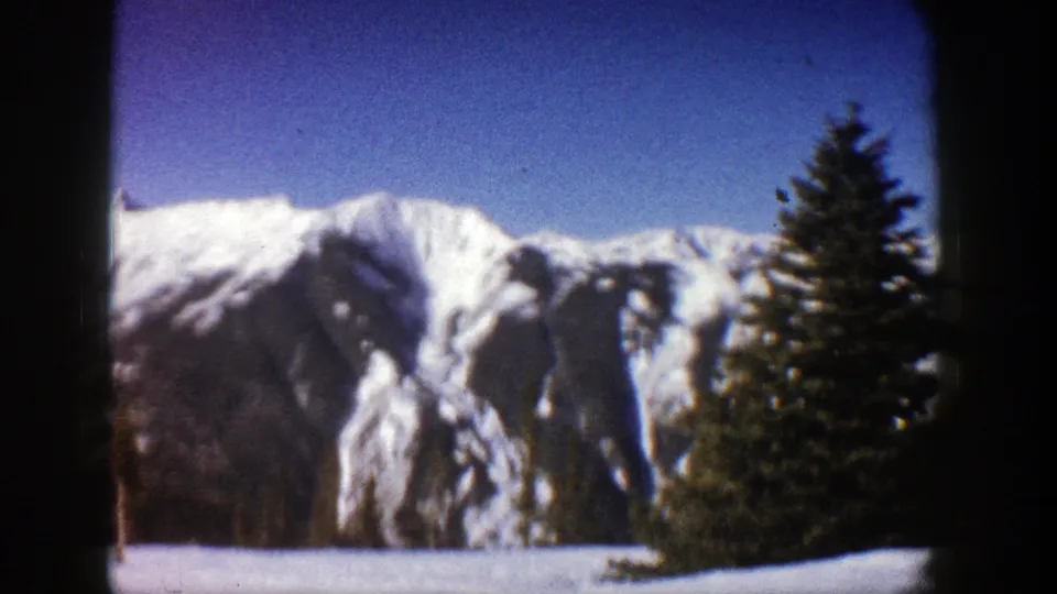 1961 Aspen, Colorado frame with snowy mountain slopes, a prominent evergreen, blue sky, and home-movie film texture.
