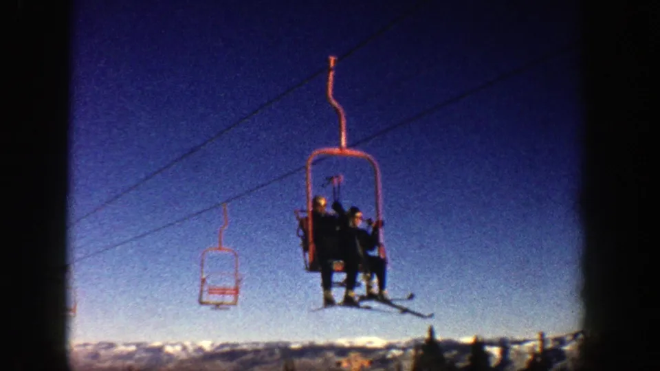 1961 Aspen ski-lift frame showing people riding an open red chairlift against a blue sky, with snow and treetops below.