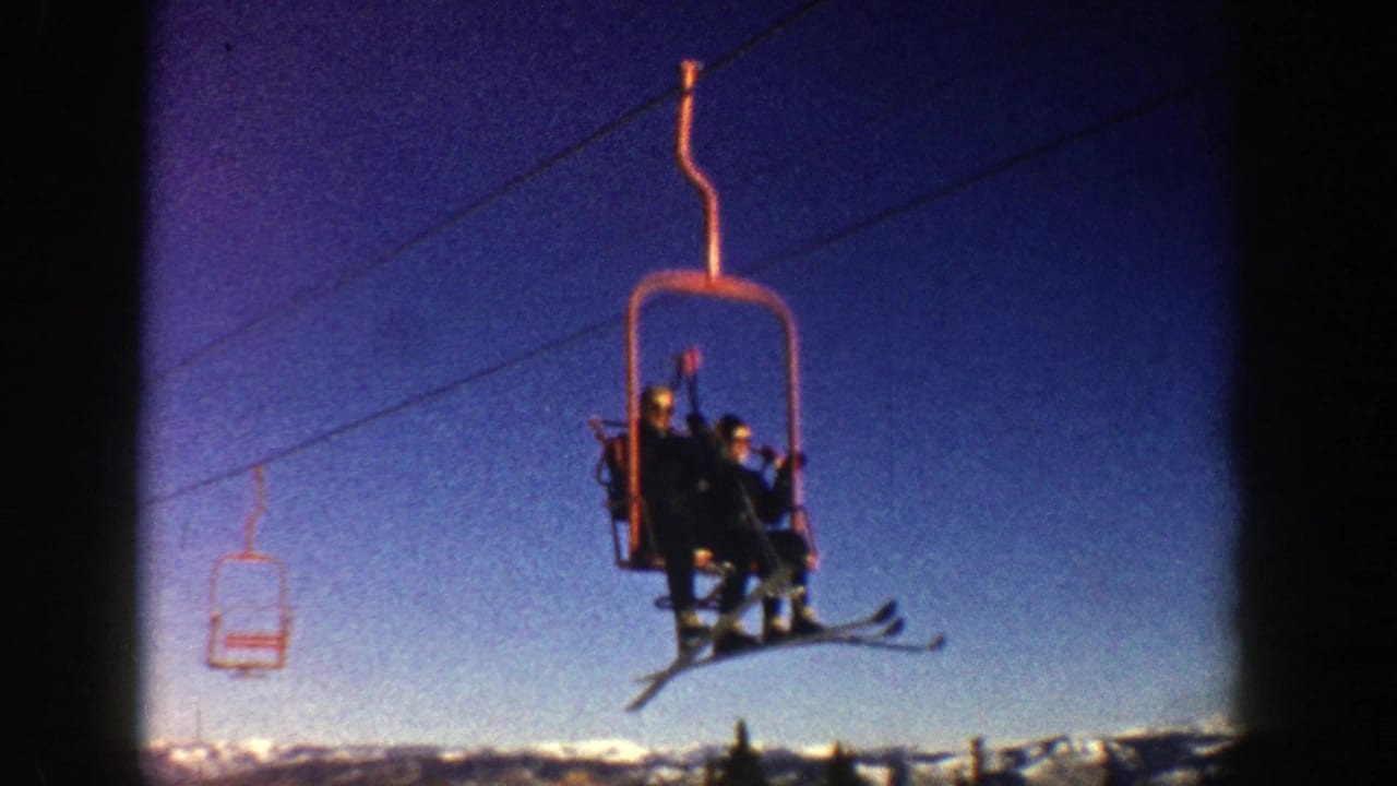 In Aspen, Colorado in 1961, two bundled skiers ride an open red chairlift with their skis hanging below against a blue winter sky.