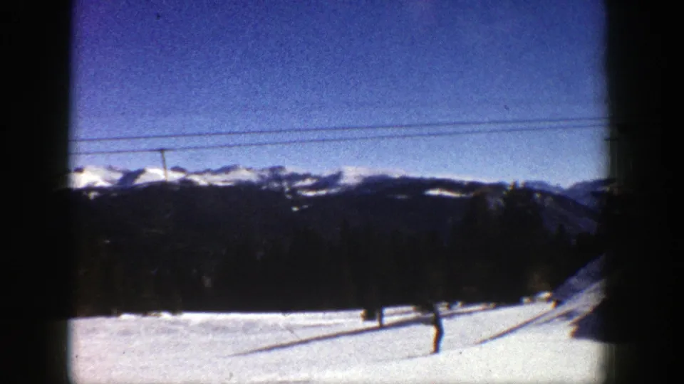 A wide 1961 Aspen ski-resort view shows a snowfield, a small skier or visitor figure, lift cables, dark forest, and distant mountains in soft 8mm color.