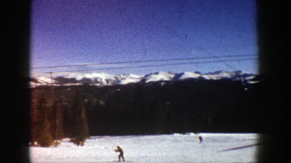 1961 Aspen home-movie frame showing a skier on a bright slope below a lift line, framed by trees and snow-covered mountains.