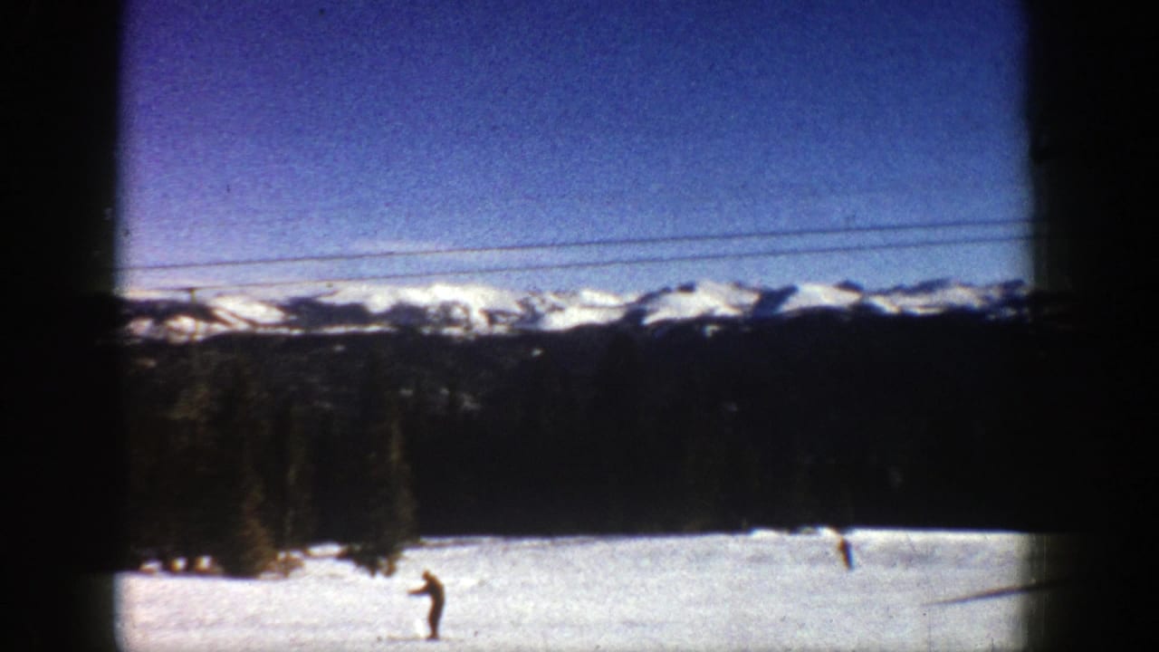 Snowy open area with small figures, dark trees, and snow-capped mountains under blue sky.